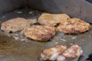 Mehrere flache, panierte Schnitzel und kleine Fleischstücke braten zischend in heißem Fett auf einer großen Grillplatte.