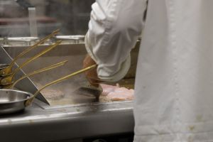 Ein Koch in weißer Kleidung wendet mit einem Pfannenwender Fleischstücke auf einer heißen Grillplatte.