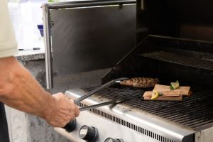 Eine Person wendet mit einer langen Zange ein Steak auf einem Gasgrill, daneben garen zwei gefüllte Holzröllchen.
