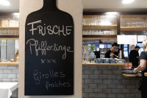 Eine schwarze Schiefertafel an einer Wand mit einer handgeschriebenen Ankündigung für frische Pfifferlinge, im Hintergrund sieht man das Personal an der Bar.