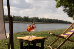 Zwei Spritz-Cocktails auf einer Kiste neben einem Liegestuhl, mit Blick auf einen Fluss an einem bewölkten Tag.