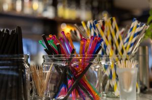 Viele bunte Rührstäbchen aus Plastik, schwarze Trinkhalme und Papierstrohhalme stehen bereitgelegt in verschiedenen Gläsern auf einer Theke.