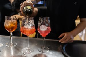 Ein Barkeeper gießt eine klare Flüssigkeit in zwei mit Eis und rötlichem Likör gefüllte Gläser ein, um Spritz-Cocktails zu mixen.