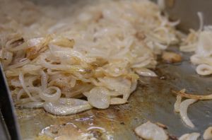 Viele in Ringe geschnittene Zwiebeln braten glasig auf einer heißen Grillplatte.