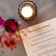 Gedruckte Menükarte mit dem Titel „Menü Womo - Dinner für 2 Personen“ auf einem Tisch neben einem brennenden Teelicht.