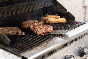 Steaks und zwei Holzröllchen garen rauchend auf einem Gasgrill, während ein Pfannenwender unter ein Steak geschoben wird.