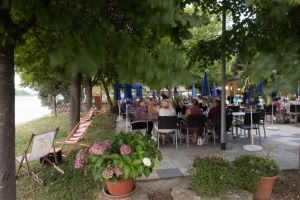 Viele Gäste sitzen gemütlich an Tischen mit blauen Sonnenschirmen auf der gepflasterten Außenterrasse des Restaurants direkt in Ufernähe.