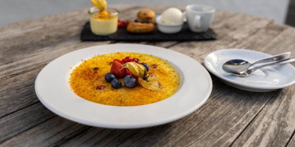 Eine große Portion Crème brûlée, garniert mit frischen Beeren und einer Physalis, im Hintergrund ein weiteres Dessert-Arrangement.