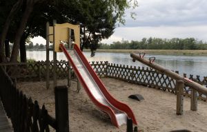 Ein Kinderspielplatz mit roter Rutsche und Holzwippe direkt am Flussufer, eingezäunt durch einen kleinen Holzzaun.