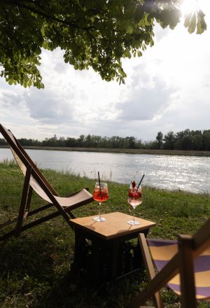 Zwei Spritz-Cocktails auf einer Kiste zwischen zwei Liegestühlen am Ufer eines Flusses unter Bäumen im sonnigen Gegenlicht.