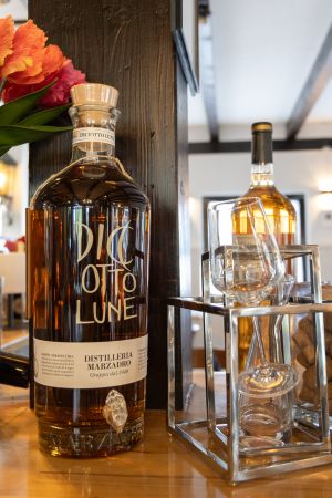 Eine dekorative Großflasche Grappa mit Zapfhahn, daneben leere Verkostungsgläser in einem Metallhalter.