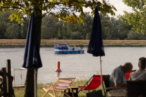 Blick durch blaue Sonnenschirme und entspannende Gäste im Vordergrund auf ein kleines Boot auf dem Fluss.