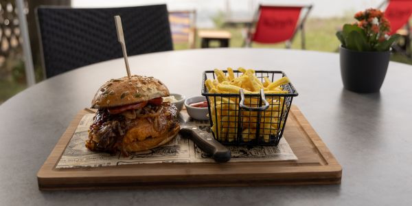 Ein Burger und eine Portion Pommes frites in einem Gitterkorb auf einem Holzbrett vor einer Flusskulisse.