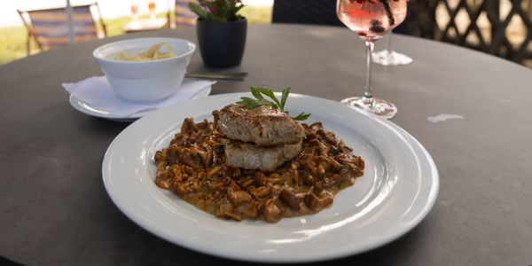 Ein Teller mit Schweinemedaillons auf Pilzrahmsoße, im Hintergrund eine Schale mit Nudeln und zwei Cocktails auf einem Tisch.