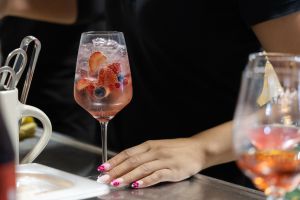 Eine Person bereitet einen rosafarbenen Sommercocktail mit Eiswürfeln, frischen Erdbeeren und Blaubeeren in einem Weinglas zu.