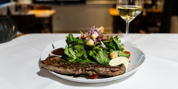 Rinderhüftsteak mit Kräuterbutter, serviert mit einem Feldsalat samt Kirschtomaten und Croûtons neben einem Glas Weißwein.