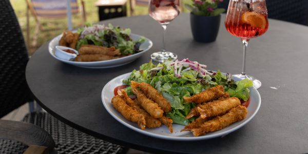 Zwei große Teller mit frischem gemischtem Salat und knusprig panierten Garnelen sowie zwei Cocktails auf einem grauen runden Tisch im Freien.