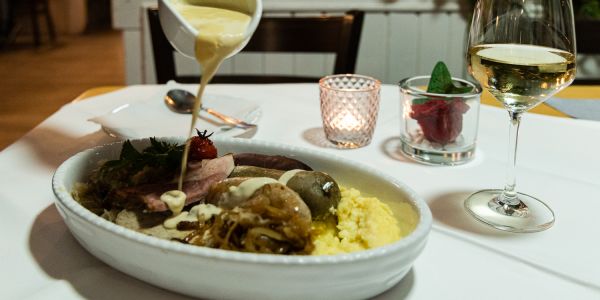 Aus einer kleinen Sauciere wird am gedeckten Tisch helle Sauce über Würste, Fleisch, Sauerkraut und Kartoffelpüree in einer Auflaufform gegossen.