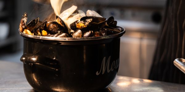 Eine cremige helle Sauce wird aus einer Kasserolle über dampfende Miesmuscheln in einem schwarzen Emailletopf gegossen.