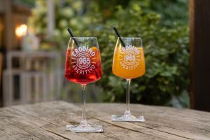 Zwei Gläser mit rötlichem und orangefarbenem Crodino Aperitivo stehen auf einem Holztisch im Freien.