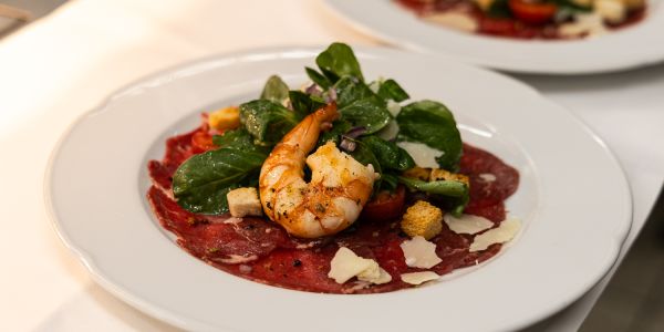 Rinder-Carpaccio auf einem weißen Teller, garniert mit einer gebratenen Riesengarnele, Feldsalat, gehobeltem Parmesan und Croûtons.