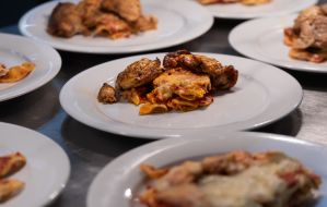 Mehrere weiße Teller mit zubereiteten Hähnchenteilen und überbackener Pasta, vorbereitet auf einer Edelstahlanrichte.