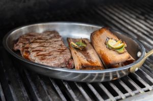 Ein gegrilltes Steak und zwei Holzröllchen in einer Edelstahlschale direkt auf einem Grillrost.