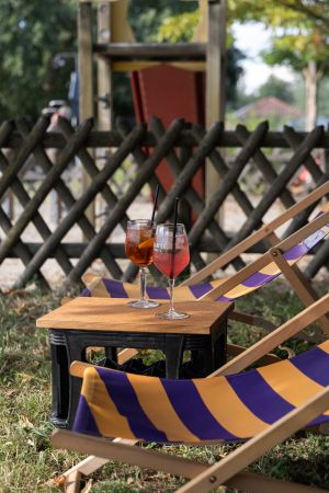 Zwei rötliche Spritz-Cocktails stehen auf einer umgedrehten schwarzen Getränkekiste zwischen zwei lila-gelb gestreiften Liegestühlen.