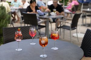 Vier bunte Spritz-Cocktails stehen auf einem Tisch auf einer belebten Außenterrasse mit Gästen im Hintergrund.