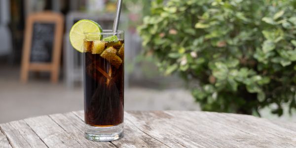 Ein Longdrink auf Cola-Basis im hohen Glas mit Eiswürfeln, einer Limettenscheibe und einem Metallstrohhalm auf einem Holztisch.
