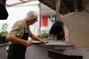 Ein Mann mit Schürze bedient den traditionellen Holzofen mit einem langen Schieber auf einer Außenterrasse.